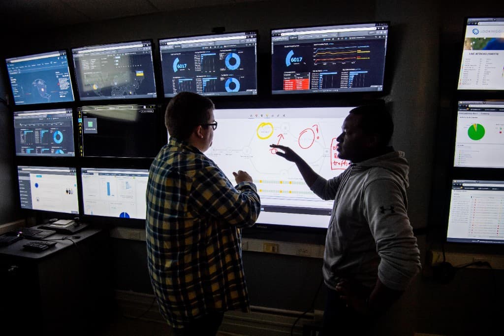 Two people review dashboards and a diagram on a wall of monitors.