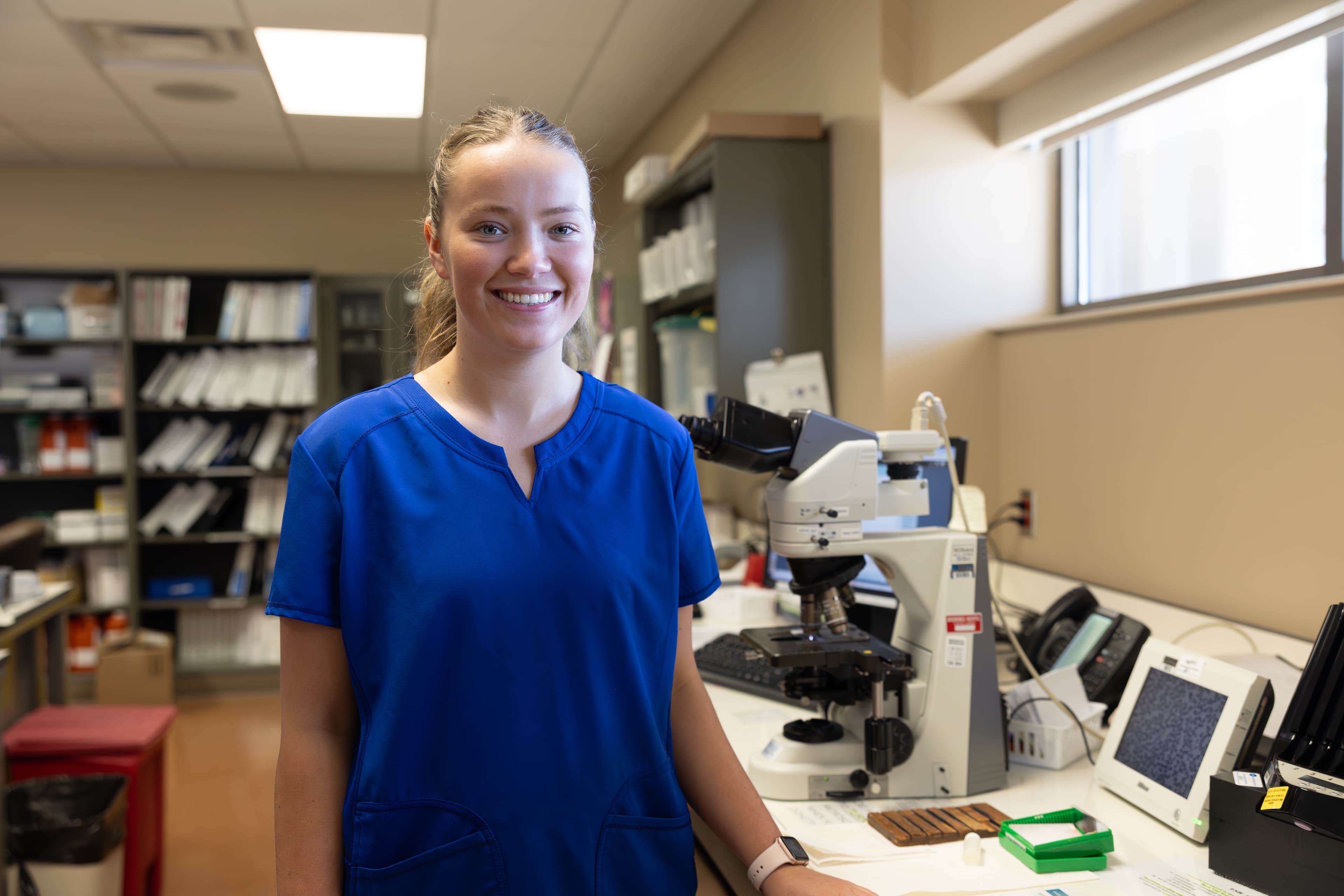 A woman in blue scrubs stands beside a microscope in a laboratory.