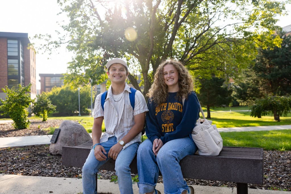 Two students sit together on a bench outside a campus building.